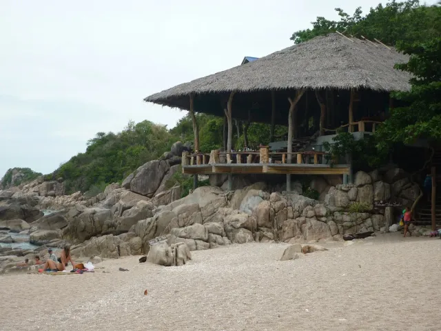 A thatched pavilion on stilts above large boulders, a sandy beach and bathers