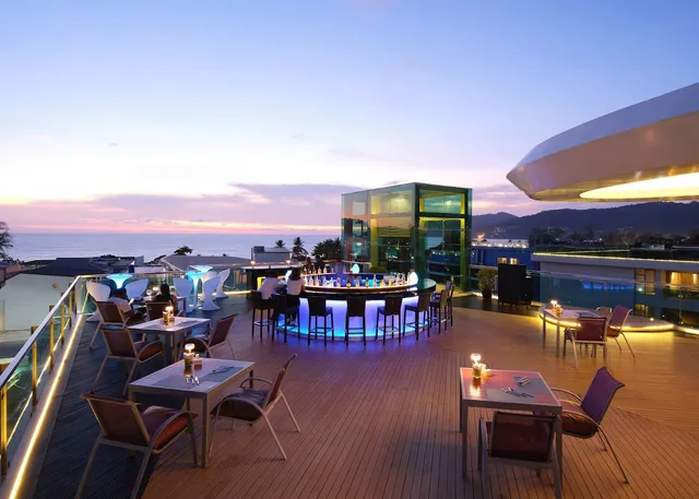 A roof terrace with candlelit tables and a blue-lit bar, facing the sea at sunset