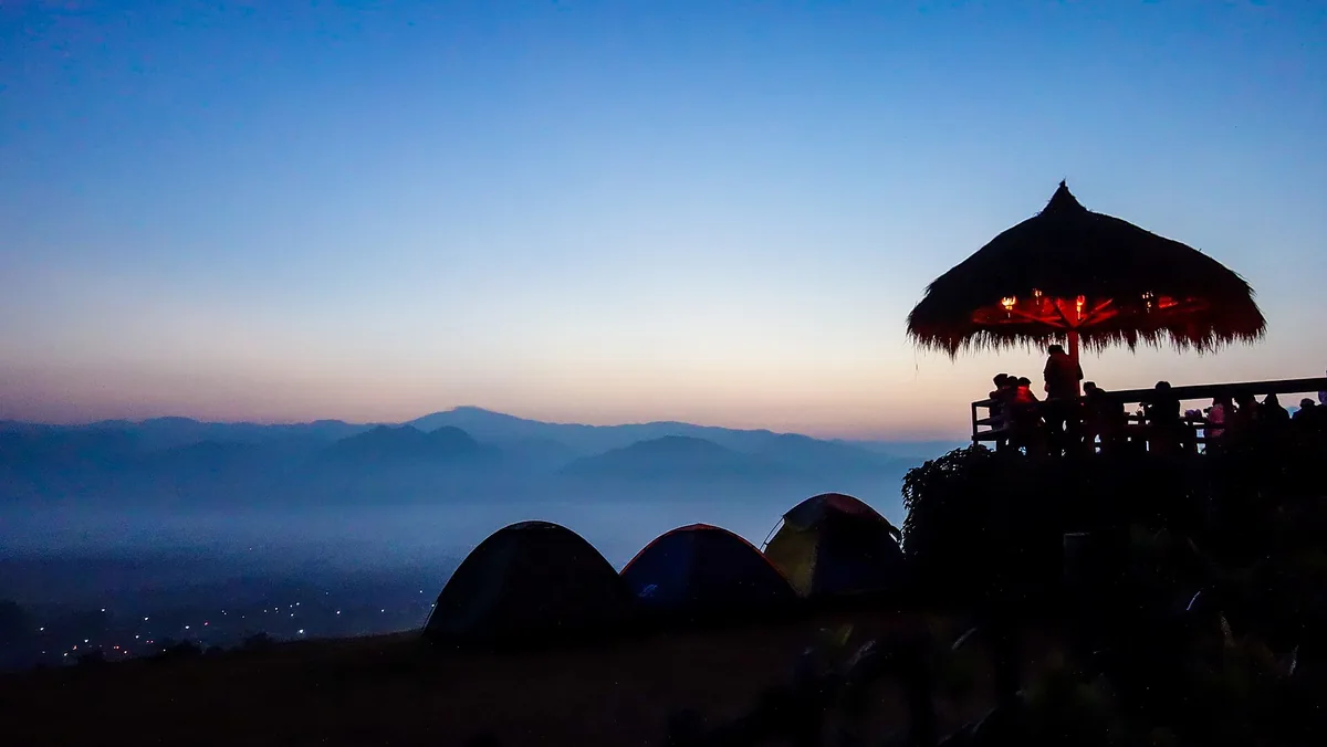 Mist wrapped mountain ridges at dawn, with tents and a viewing hut silhouetted on the summit