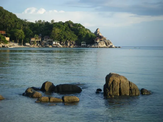 Rocks poking out of calm water and wooden bungalows on a forested slope behind