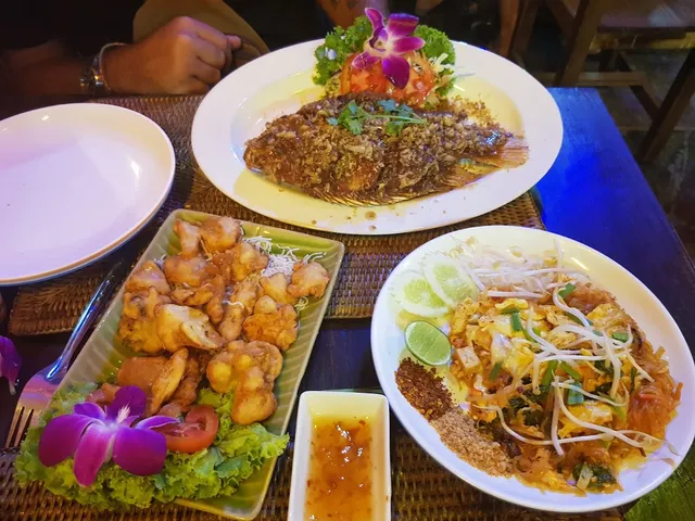 Fried fish, pad thai and a fried dish on a table, decorated with purple orchids