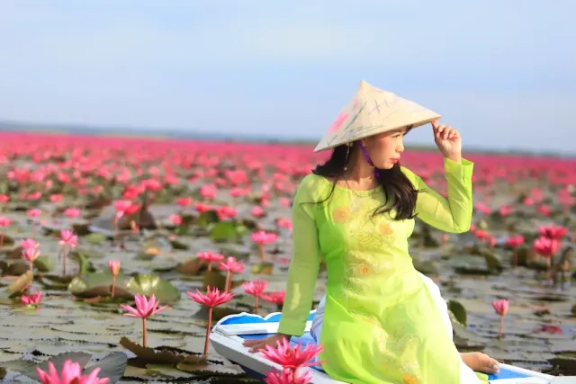 A woman in a green dress and conical straw hat sitting in a boat in a pink lotus field