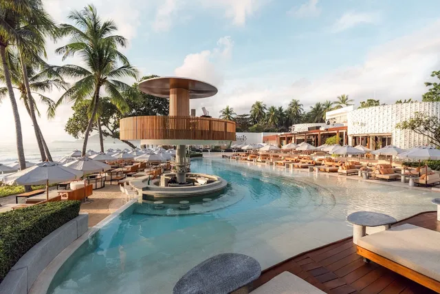 A curving pool with a raised round bar at its center, palms and parasols along the beach