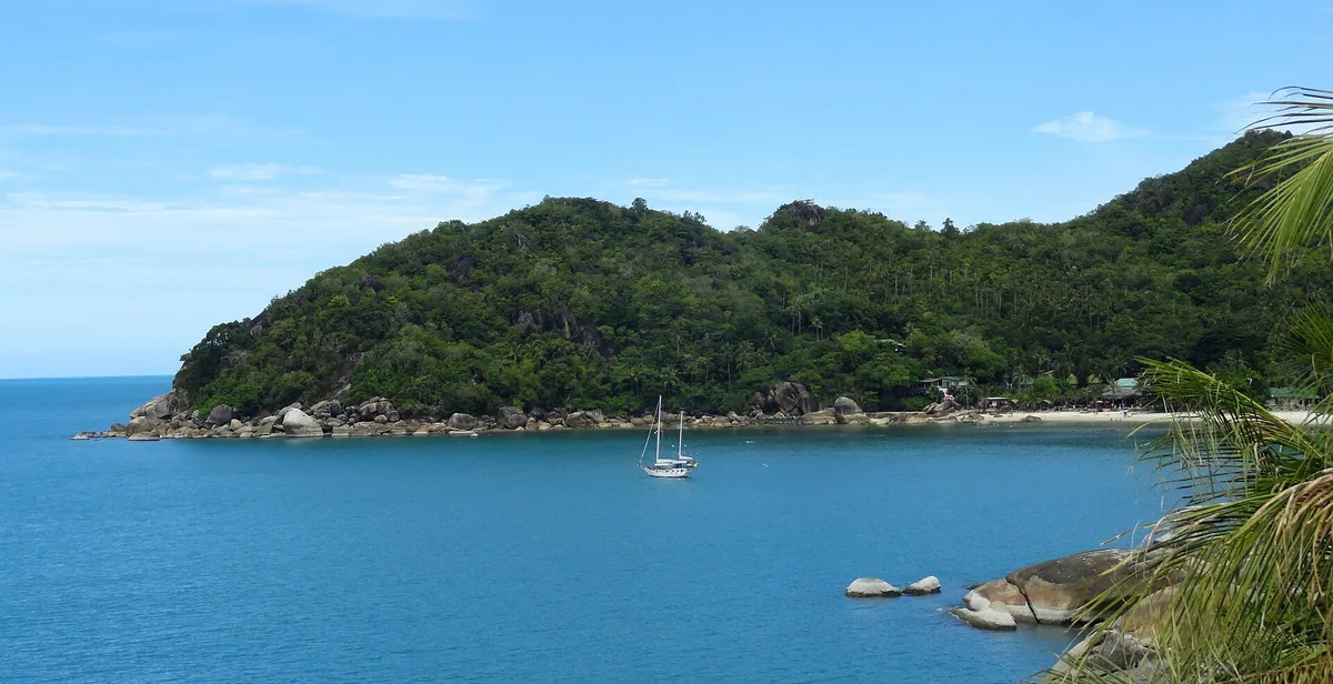 A small turquoise bay with a single sailing boat, framed by a green hill and granite boulders
