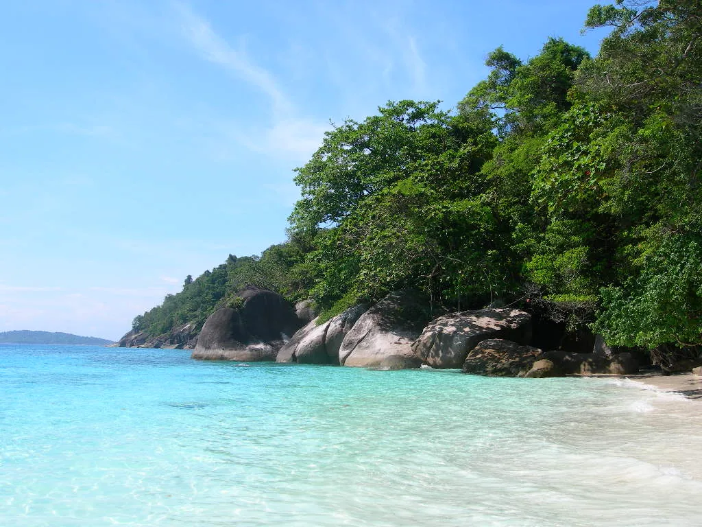 Clear turquoise water on a white sand beach, with granite boulders and green jungle along the shore