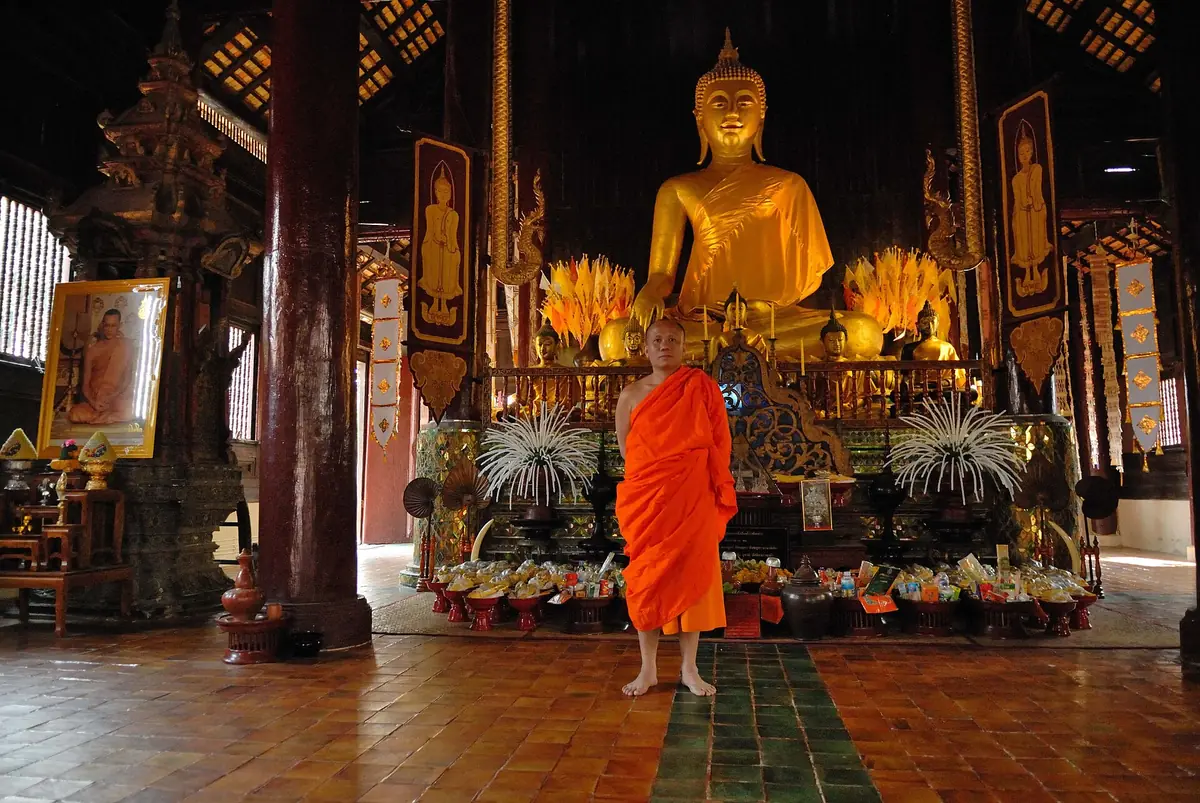 Monk in an orange robe standing in a teak hall before a gilded Buddha and candles