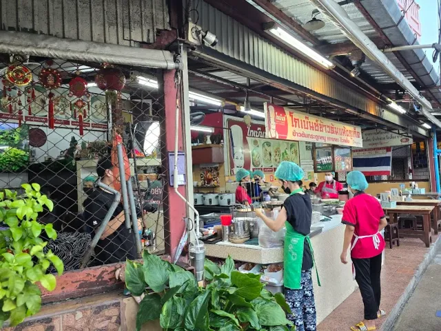 A roadside cooking stall with workers in aprons and hairnets beside stainless steel pots