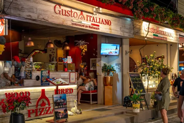 A lit front at night with a red sign, a wood fired pizza sign and passersby