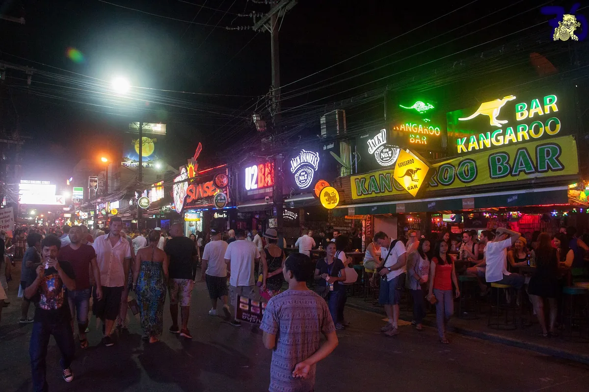A bar street at night with green and red neon signs and a large crowd on the road