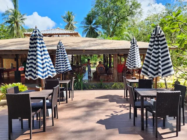 A deck with blue and white striped parasols, rattan chairs and a thatched roof building