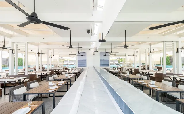 A bright white dining hall with wooden tables and ceiling fans, and a pool beyond the windows