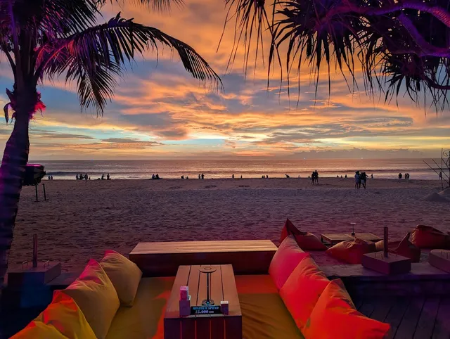 Sofas with orange cushions on a wooden deck facing a wide beach at a colorful sunset
