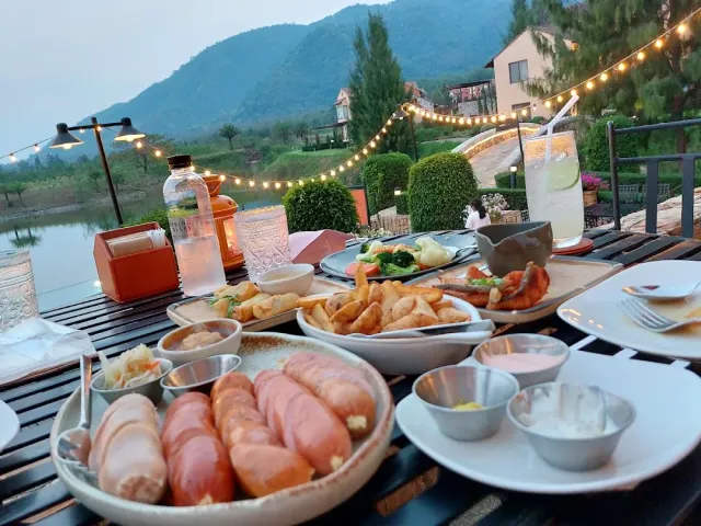 A table loaded with sausages, fries and sauces facing a lake and hills at dusk