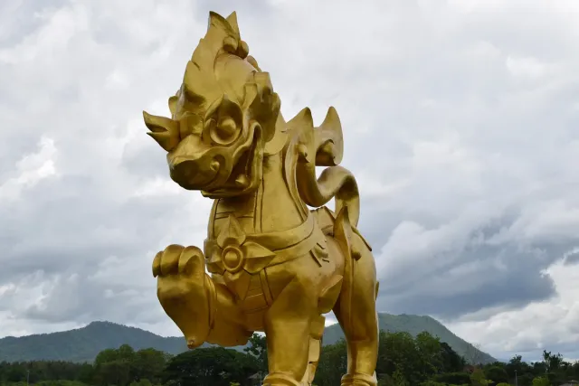 A huge gilded lion statue standing against green fields and a cloudy sky