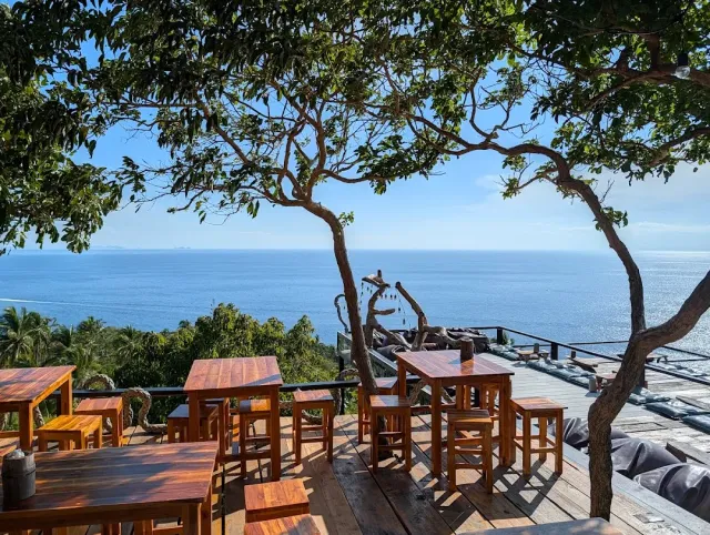 Wooden tables on a deck above the sea, tree branches spreading shade over them
