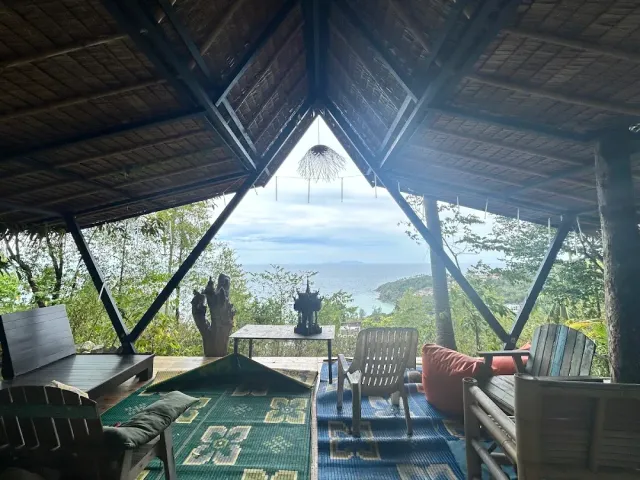 A triangular pavilion with a thatched roof, mats and cushions and a bay view between the trees