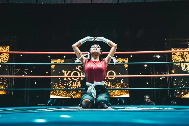 A fighter kneels in a boxing ring with raised arms, against a gold lit backdrop