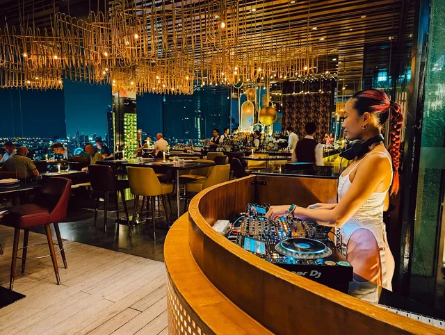 A DJ at a curved mixing booth, with diners' tables and city lights in the windows behind her