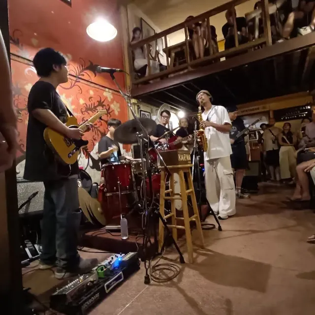 A band playing on a small stage, guitar, drums and saxophone, and an audience on the balcony