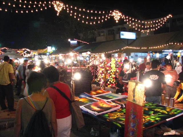 Night market stalls loaded with goods under strings of bulbs, with shoppers browsing between them
