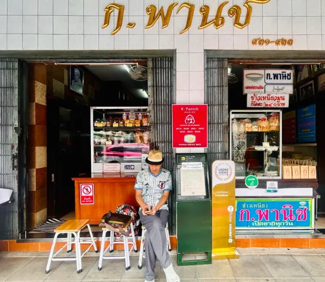 A shopfront with golden Thai letters, display cases and a man sitting at the entrance
