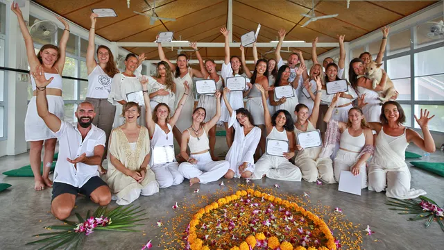 A group of graduates in white holds certificates in a practice hall, around a flower mandala on the floor