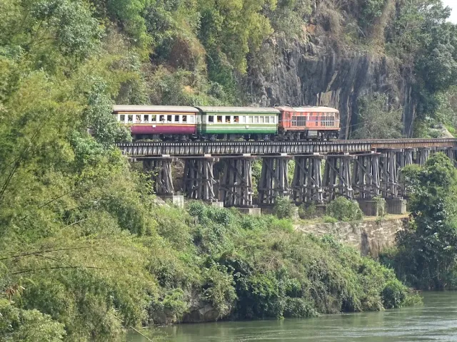 A passenger train crossing a wooden bridge hugging a cliff above a greenish river