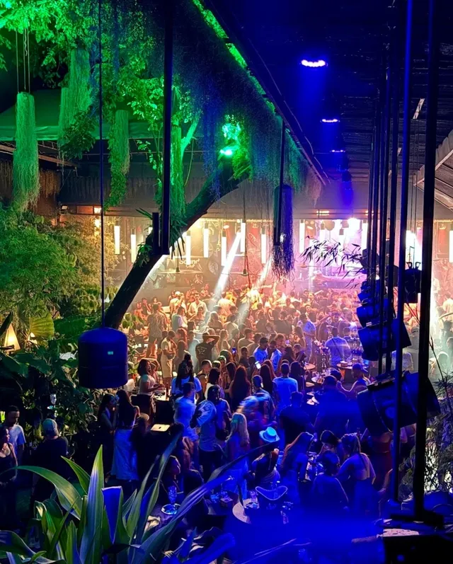 A crowded dance floor among hanging greenery, with bright stage lights in the background