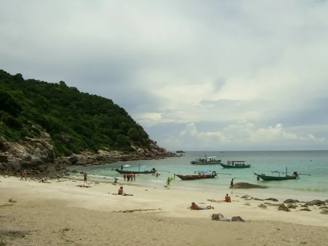 Longtail boats anchored off a beach with bathers and a green hill above