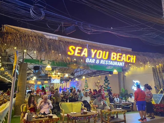 A thatched roof bar and restaurant with a lit neon sign and diners in the evening