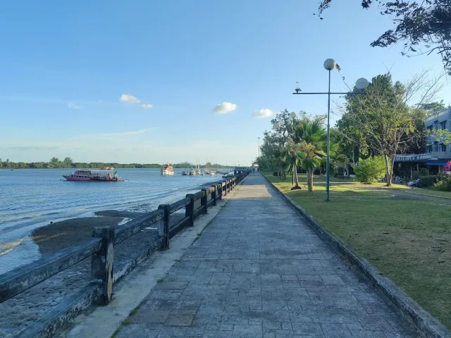 A stone promenade along the river with a railing, lamps, grass and boats in the distance