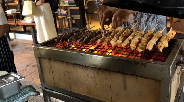 Meat skewers on a glowing charcoal grill, hands turning them over the embers
