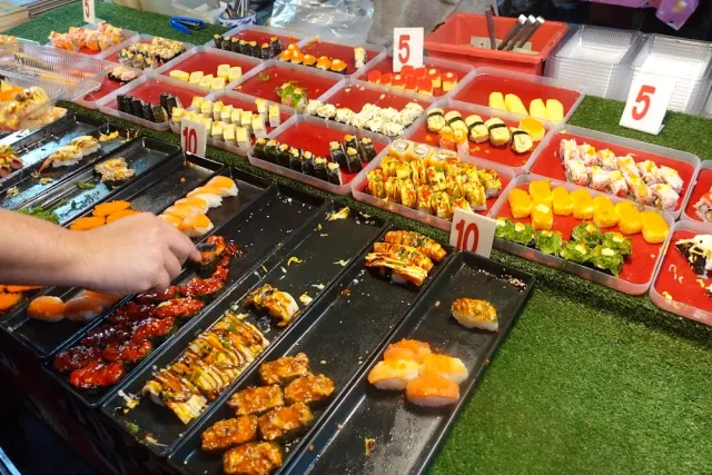 A market stall loaded with colorful sushi trays and price signs, a hand arranging a portion