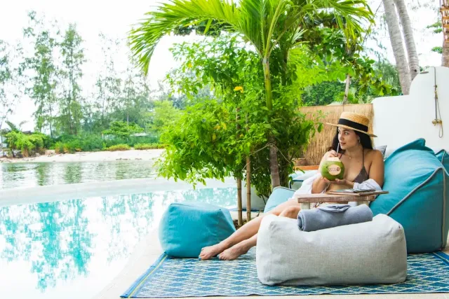 A woman drinking from a coconut on a beanbag beside a pool, a beach and palm trees behind