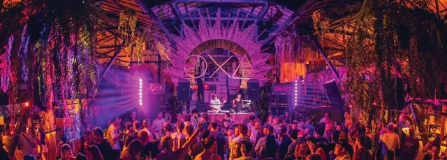 A dense crowd dancing under a woven canopy in purple light, with a lit DJ booth
