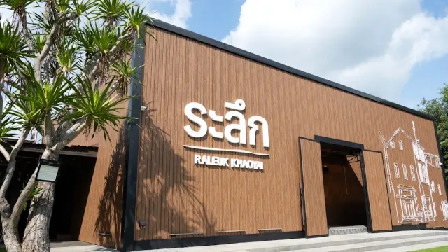 A rectangular wood clad building with a raised sign, a dark entrance and a palm to the side