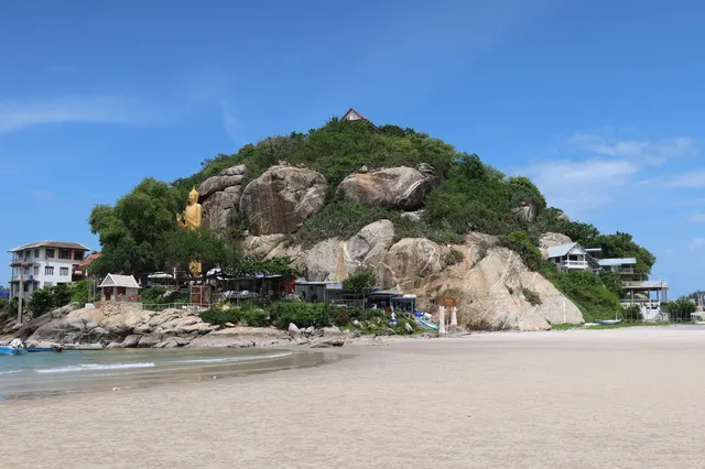 A green rocky hill with a large golden Buddha statue above a wide beach