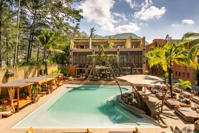 A large pool surrounded by sun beds and cabanas, palms and a concrete building behind