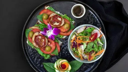 A dark plate with two slices of bread, tomato, an orchid flower and a bowl of salad