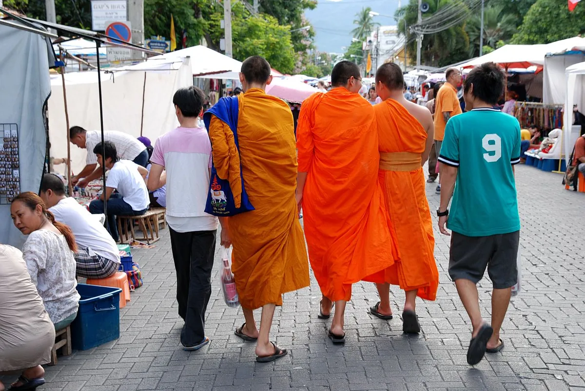 Monks in orange robes walking through a paved street market, between stalls and white canopies