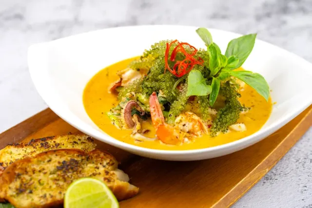 A white bowl of yellow curry with shrimp, sea grapes and basil, beside garlic bread