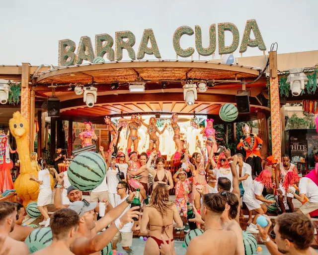 A crowded pool party beneath a giant letter sign, with costumed dancers on the stage