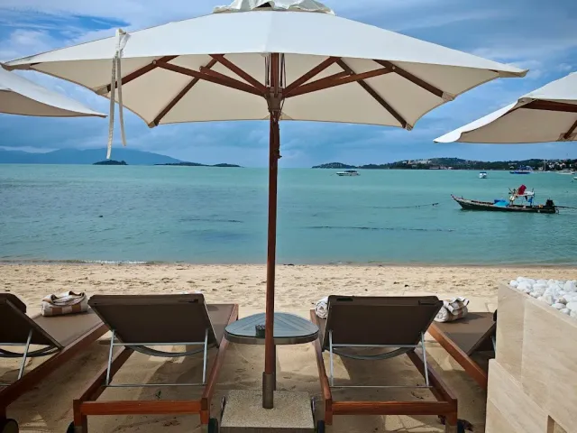 Two sun loungers under a white parasol facing calm water and a longtail boat