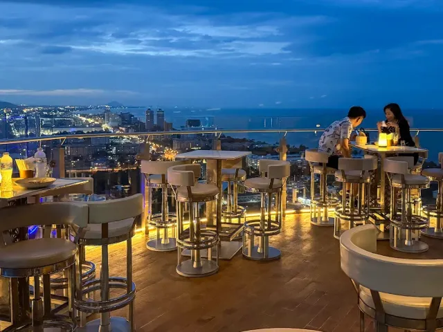 A rooftop bar at dusk with high tables, facing the city lights and the sea