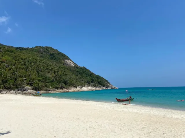 A wide white sand beach with a forested hill at its end and a longtail boat