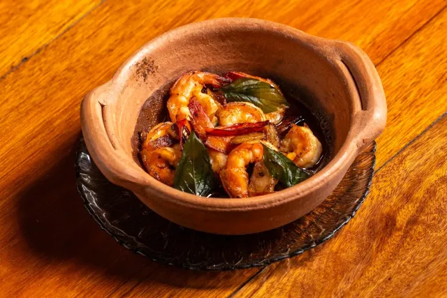 A clay bowl with shrimp, chili and green leaves on a glossy wooden table