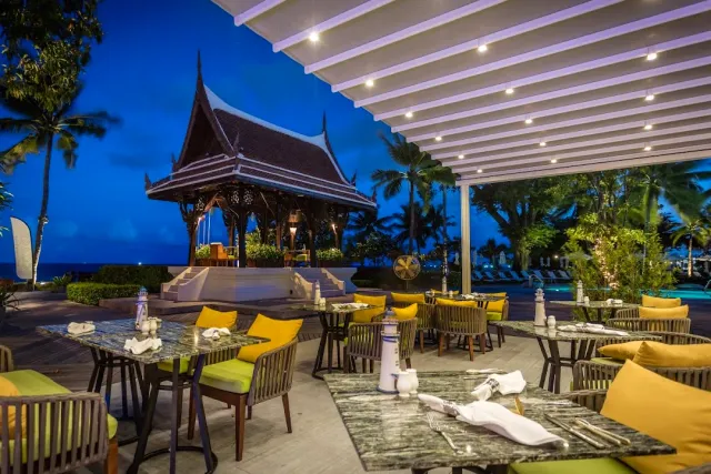 An evening terrace with yellow cushions, a roofed Thai pavilion and lit palms in the background