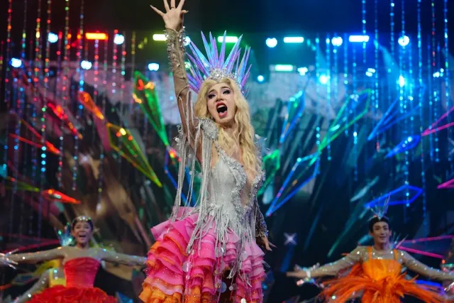 A performer in a sparkling crown and pink dress on stage against a glittering backdrop