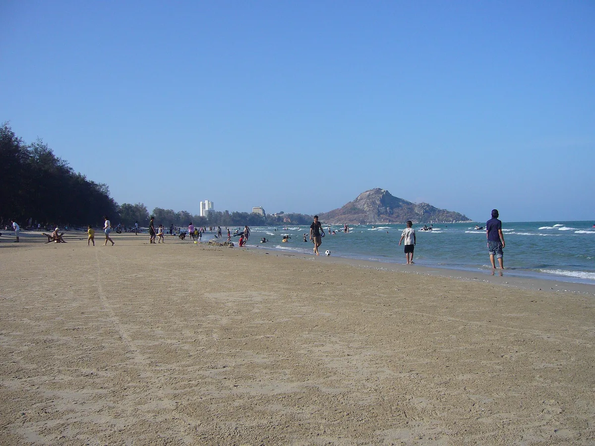 A long sandy beach with bathers in the water and a rocky headland closing the bay, on a sunny day