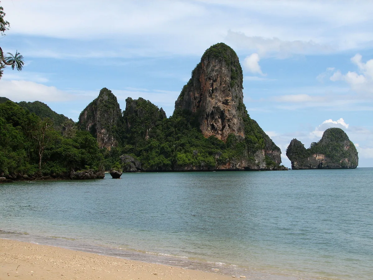 Tall limestone cliffs covered in greenery above a calm bay, with a strip of pale sand in front